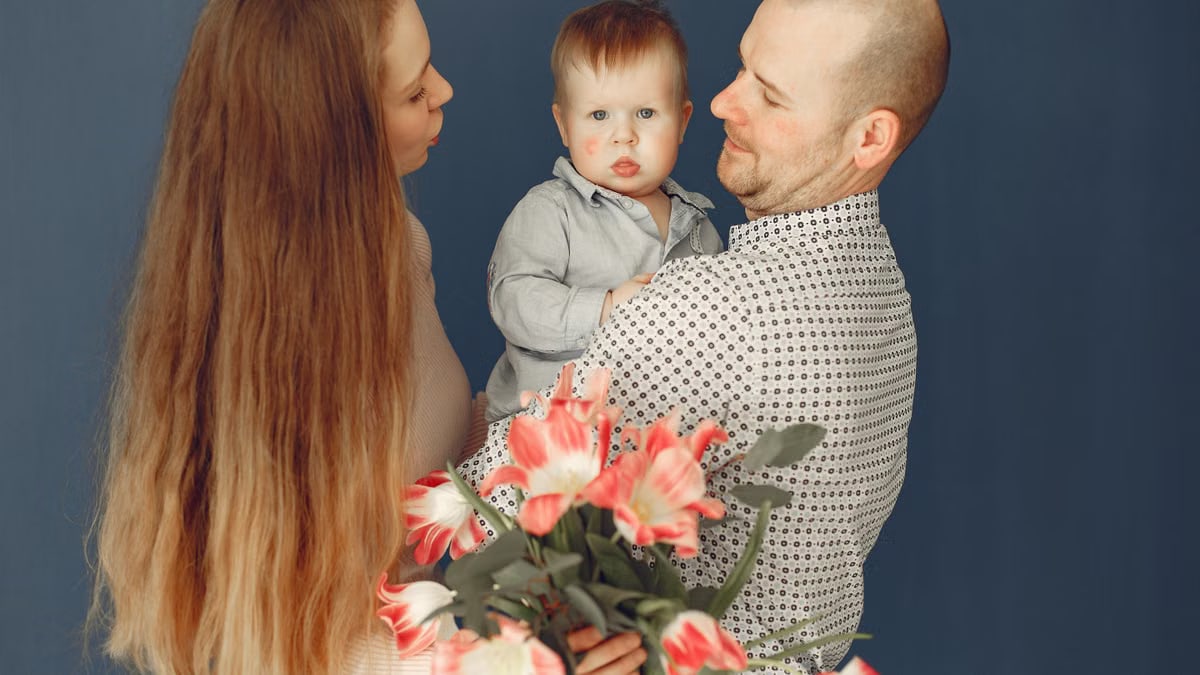 Cheerful long haired woman with bouquet of flowers embracing husband and little son while father holding boy against blue background